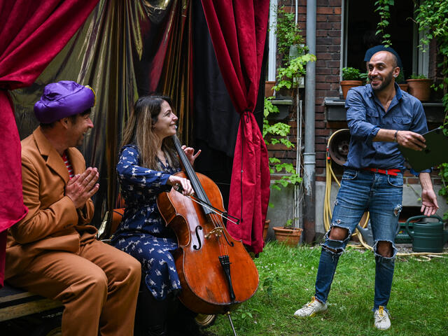 Bashar stands and talks to two of his colleagues, who are sitting on the mobile stage Bashar uses. One colleague with a blue hat is clapping his hands while the other colleague, a woman, holds a cello. 