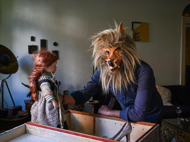 Bashar wears a wolf mask while holding a doll with red hair.