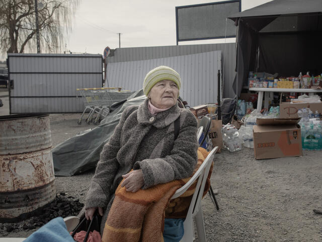 A older woman sits on a chair with a blanket and looks at the camera. There are supplies and other refugees in the background. 
