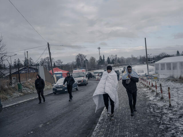 Two refugee men walk down the street with blankets over the shoulders. 