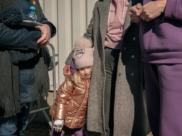 A young girl wearing a winter coat and hat leans on a woman standing next to her.