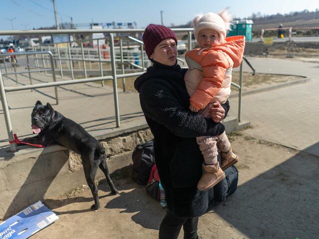 A mother carries her young daughter while speaking to the camera. A large black dog stands behind her. 