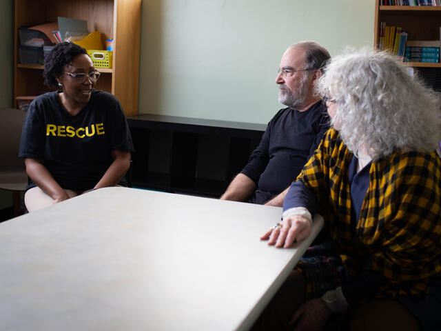 Diana Mbogoni sits at a table talking to Mandy Patinkin and Kathryn Grody. 