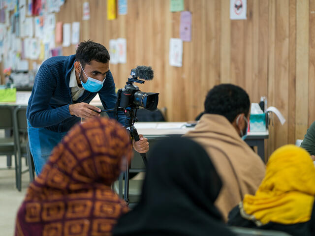 Anwar bends over and looks through his camera while Afghan refugees sit nearby listening to an IRC staff member.