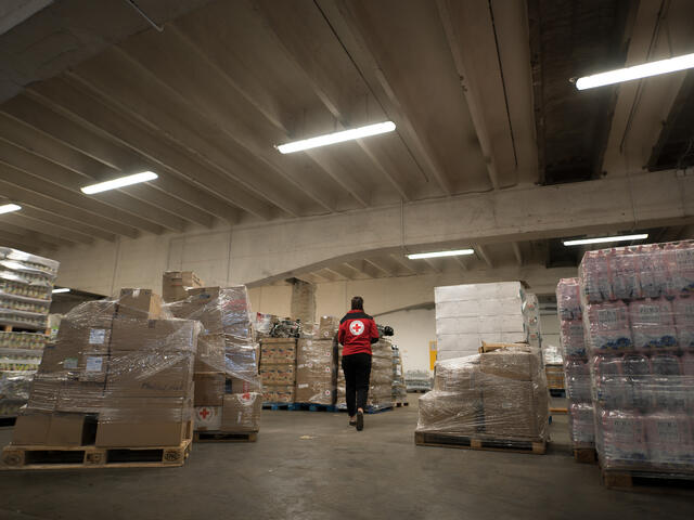 A person with a Red Cross jacket walks through a warehouse of supplies, their back to the camera.