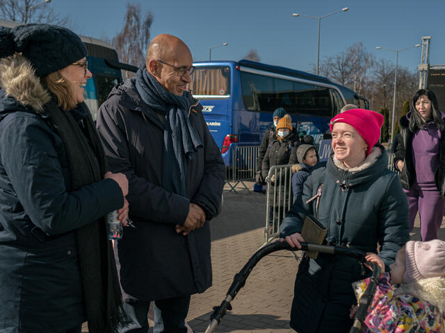 Three people stand outside in winter coats, smiling and speaking.