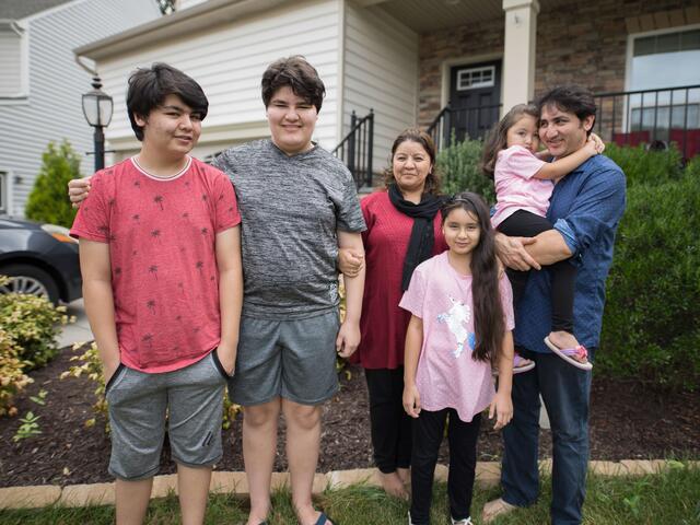 A family--husband holding a young girl, his wife, one other girl and two boys--stand outside their home. 