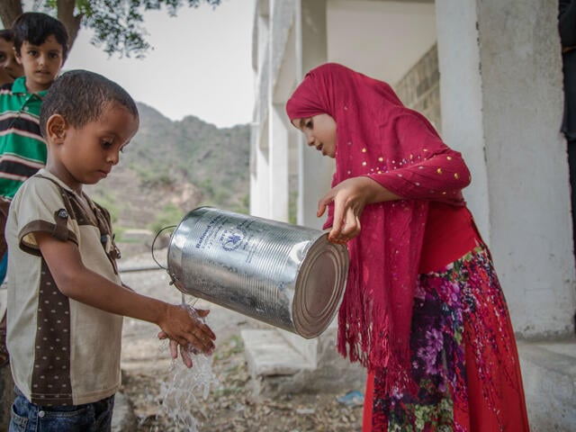 A girl  holding a jug pours water over a boy's hands so that he can wash them.