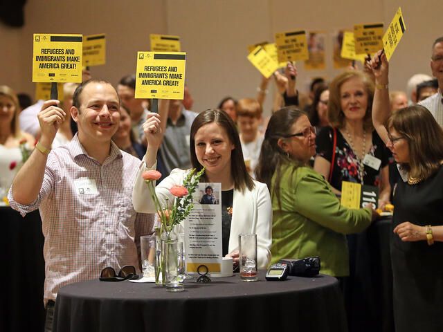 Multiple attendees at Journey to the Wasatch lift their auction paddles with smiles, signs on the paddles reading "Refugees and immigrants make America great!"