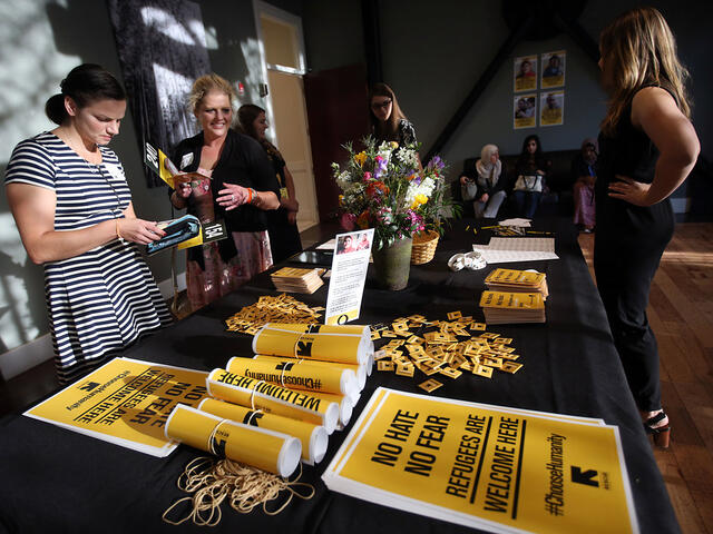 A table displaying posters reading "No hate no fear, refugees are welcome here #ChooseHumanity" and an IRC logo. The table has pins other posters and flowers, and women are examining and grapping the items.