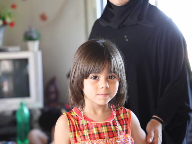 Young girl with brown hair looks at the camera. The hand of her mother rests on her shoulder.