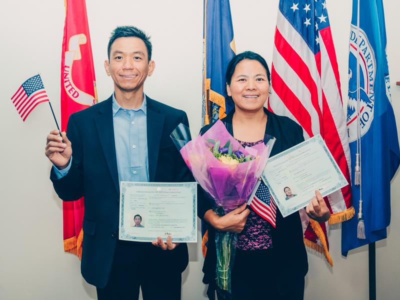 Sui and her husband Aung smile for the camera in front of large Utah and United States flags, holding small American flags, flowers, and their new naturalization papers.