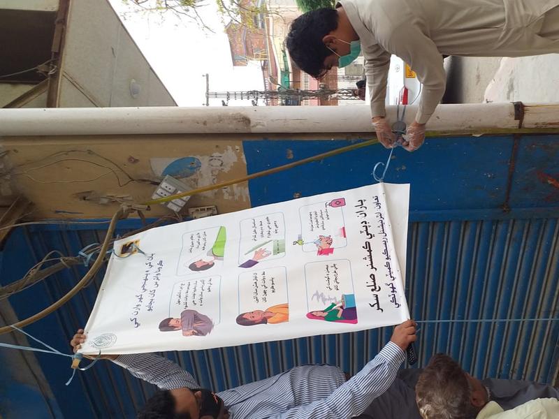 IRC educational materials are displayed in areas with high foot traffic in Sukkur, Pakistan. Two men wearing masks stand on either side of a poster with COVID-19 information.