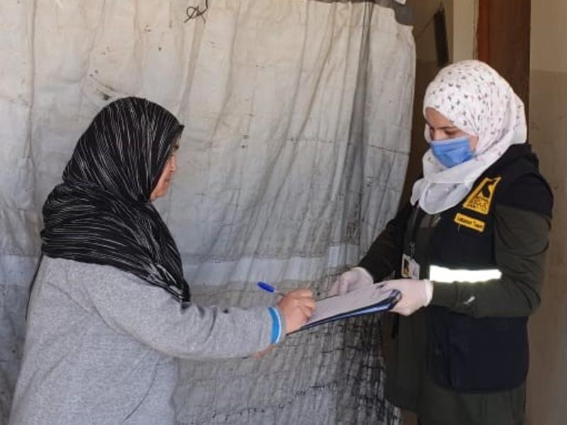 A woman in an IRC vest holds a clipboard while talking to an older woman and distributing cash assistance. 