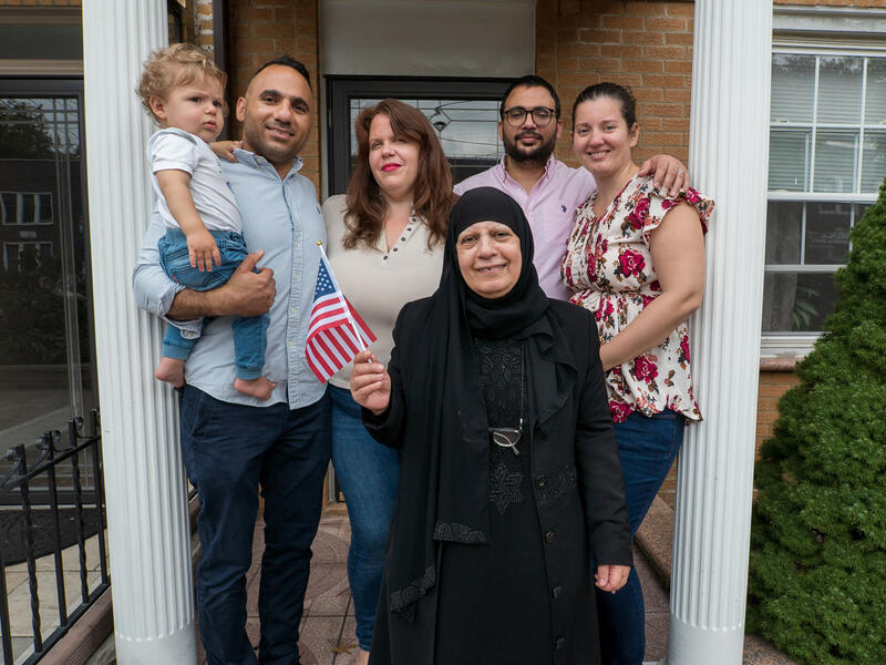 Maha and her family pose on the front porch of a house. Maha stands in front holding an American flag. Her son stands to her right holding her grandson and next to him are his wife and her other son and daughter-in-law. 