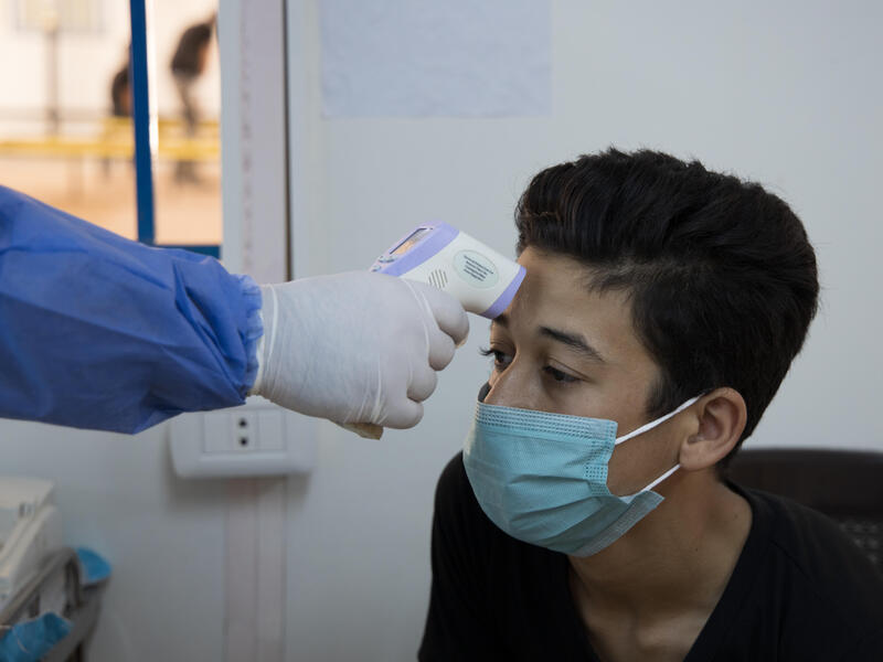Wearing a mask, Muhammad sits in a clinic while a doctor wearing full PPE takes his temperature. 