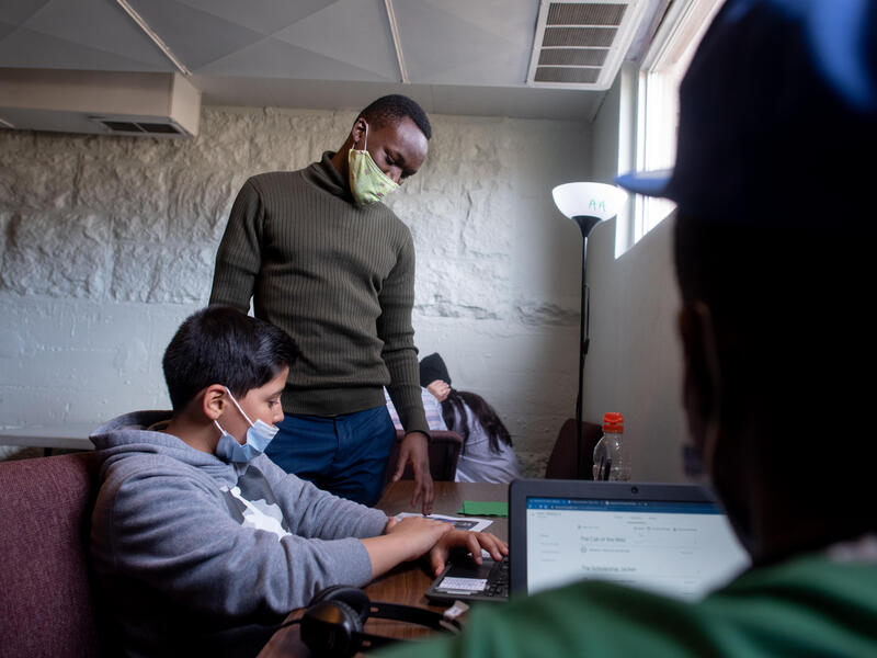 Fredrick stands next to a student at an after-school program. Both are wearing masks and the student is on his computer while Fredrick looks over his shoulder. 