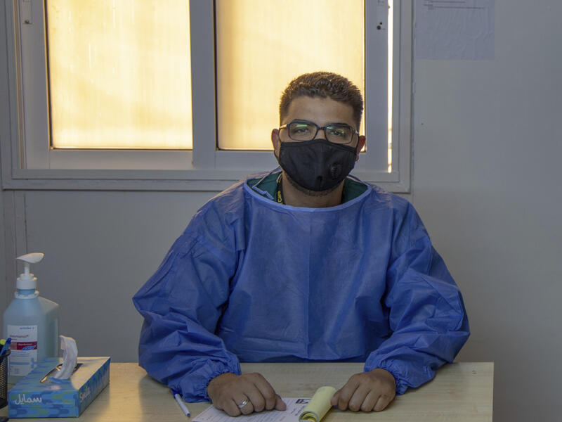 Dr. Abedalhaleem Albalasmeh, wearing a mask and a medical gown, sits behind a desk at an IRC clinic in Jordan. 