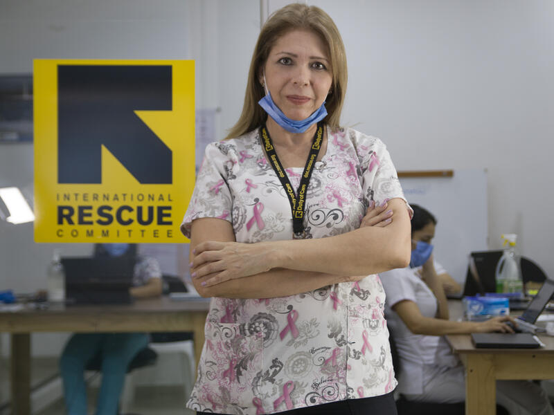 Dr. Edna Patricia Gomez stands in front of an IRC logo in a clinic wearing her medical scrubs and a mask around her chin. 