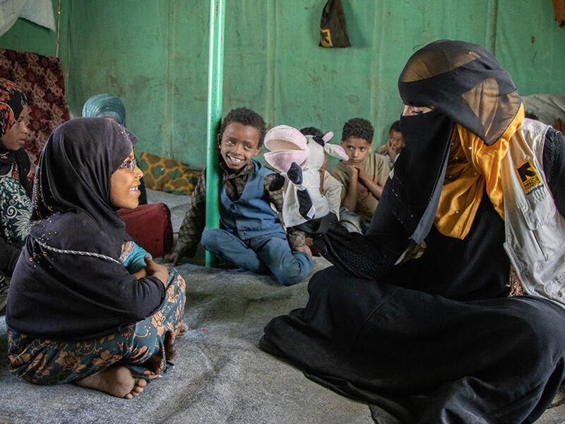 A group of young children sit while watching a puppet show. One girl interacts with the volunteer talking to her with a cow puppet