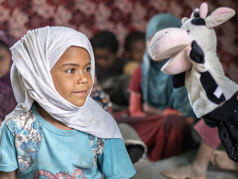 Six-year-old Ashwaq interacts with a cow puppet as part of a show in Al-Manshar camp In Al-Manshar camp, a litle girl sits and looks at a cow puppet that a volunteer is using to teach her about COVID-19 prevention.