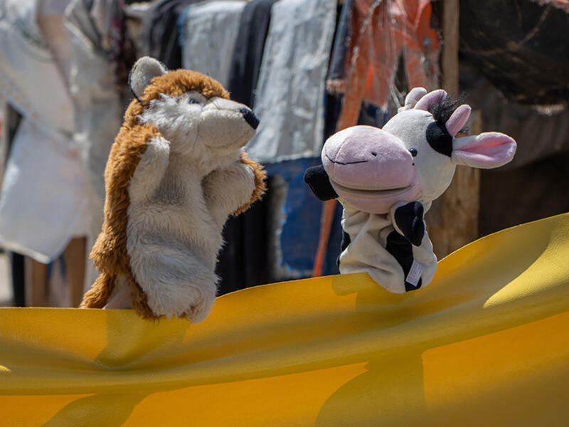 Using a yellow tarp as their "stage," a cow and fox puppet have a "conversation" during a puppet show on COVID-19 prevention 