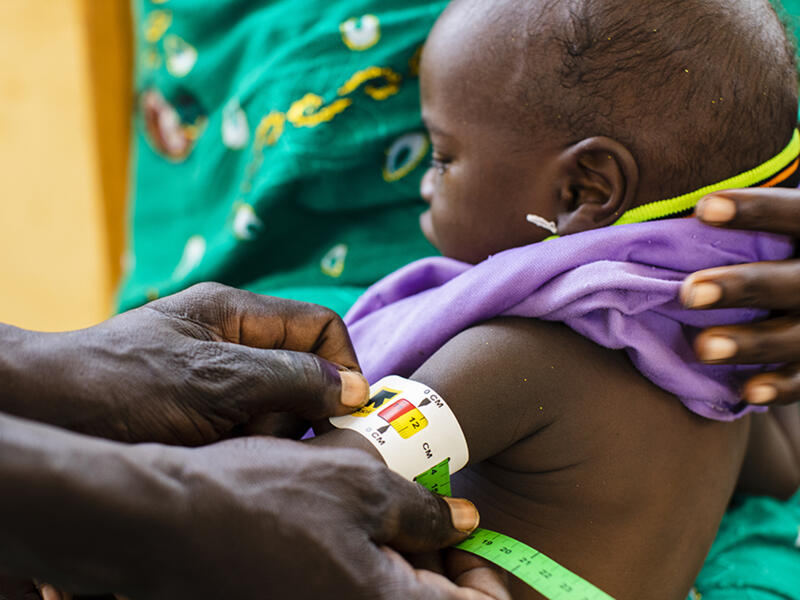 A mother, Aziza Djibrine, holds her child, Mahamat Saleh Moussa as a community health care worker uses a MUAC (middle upper arm circumfrence) band to diagnose acute malnutrition at an IRC clinic. A mom holds a small child on her lap while we see the hands of a community health worker holds a MUAC (middle upper arm circumfrence) band around his arm.