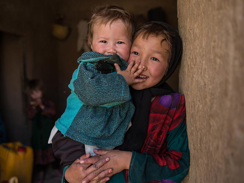 A girl holds her younger sister and looks at the camera, both are smiling and laughing. 