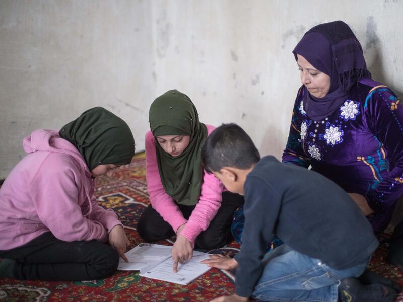 Reema with her twins Shurouq and Shirina and their brother Eissa. Reema sits on the ground with her twins Shurouq and Shirina and their brother Eissa. Their children are looking at schoolwork while Reema looks over their shoulders.