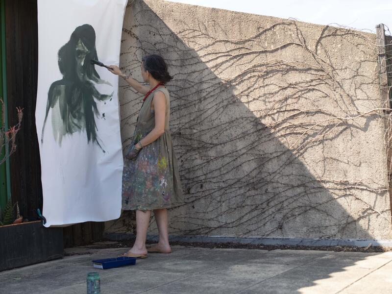 Diala Brisly, wearing a paint spattered smock dress, stads outside on a terrace painting a larger-rthan-life face on a sheet of paper hanging on the wall.
