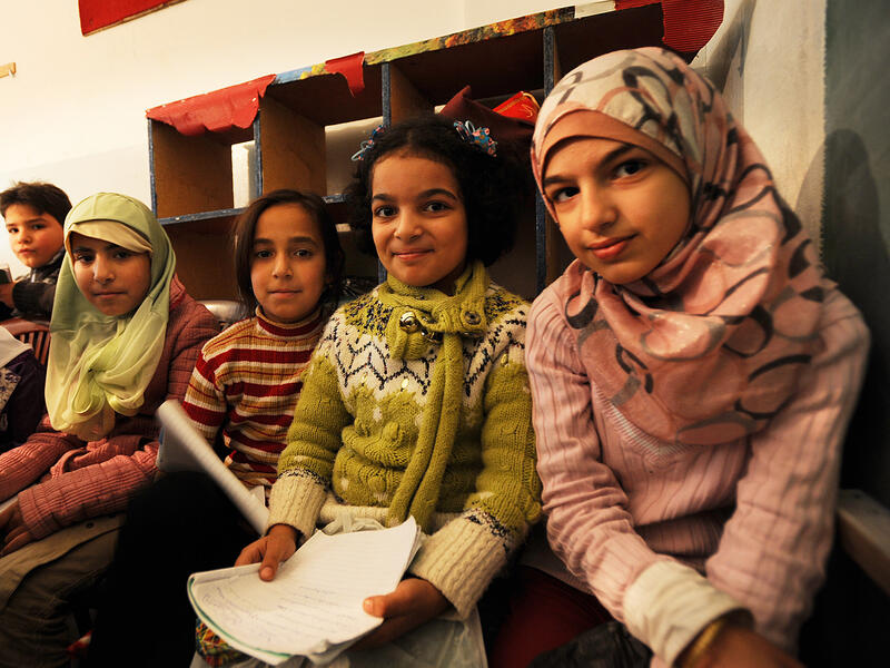Syrian refugee children in a makeshift school in Berkayel, Lebanon. Four young girls sit next to one another in a school, looking at the camera and holding school supplies.