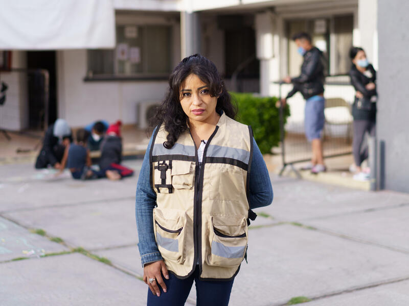 Rosa, wearing a brown vest with large pockets, stands in front of the triage hotel she helps manage in Ciudad Juárez, Mexico. In the background, children draw with chalk on the ground with their parents while other adults converse. 