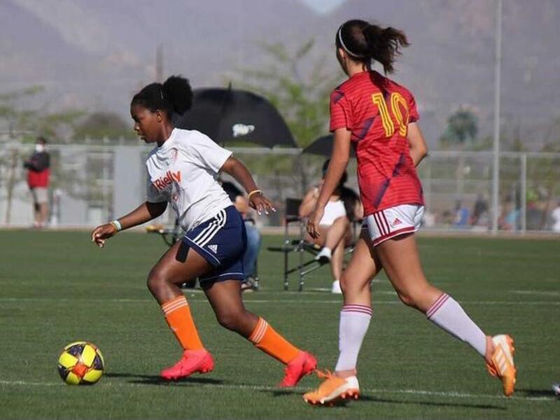 Kezia kicks a soccer ball while another player approaches her 