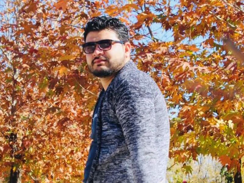 Ahmad looks at the camera while wearing sunglasses and standing in front of fall foliage. 