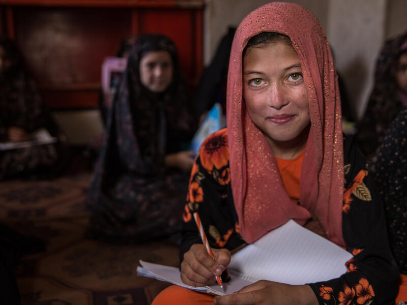 A young girl in Afghanistan at an IRC-supported Safe Healing and Learning Space. A young girl sits with a notebook on her lap ready to write while looking up at a teacher. There are other young girls sitting behind her.