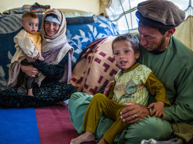 A family internally displaced in Afghanistan due to drought. The country is facing conflict, COVID-19 and the impacts of climate change. A family of four sit on rugs on the ground: a young boy sits on the lap of his father and a baby sits on the lap of her mother.