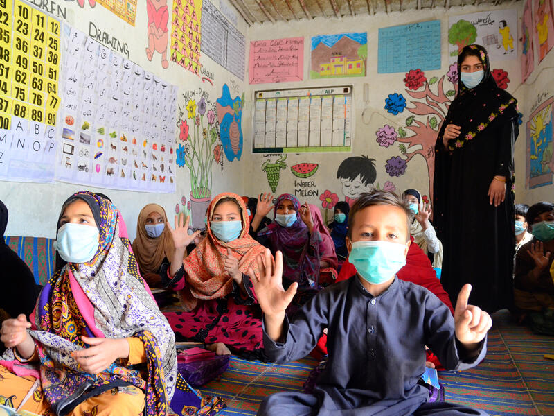 Children sit on the floor and a female teacher stands in a classroom, all are wearing masks and the kids are clapping. 