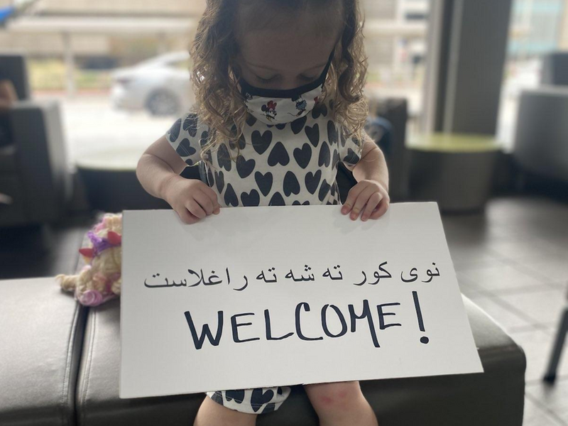 Three-year-old Juniper greets Afghan refugees at the Sacramento airport with a homemade sign in Pashto and English. A young girl holds a "Welcome" sign in Pashto and English.