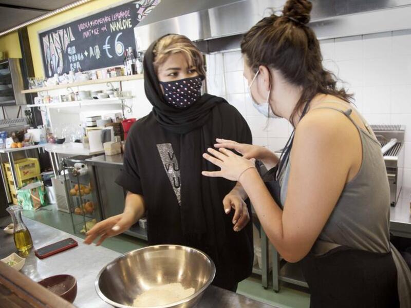 A cooking workshop with refugees on the island of Lesbos. In a kitchen, two women, both masked, talk to one another as they mix flour and spices in a large bowl.