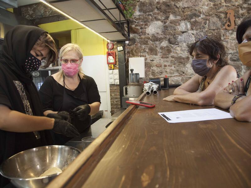 In a kitchen, Hafea stands next to one woman and shows her a technique while two additional woman sit on the other side of a counter watching. 