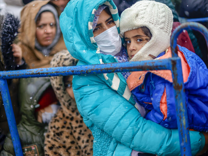 Na granicy polsko-białoruskiej w tłumie stoi kobieta w jasnoniebieskim płaszczu zimowym, trzymająca w ramionach dziecko również owinięte w zimowe ubrania.  Stoi za metalową bramą, a wszyscy na zdjęciu są stłoczeni. 