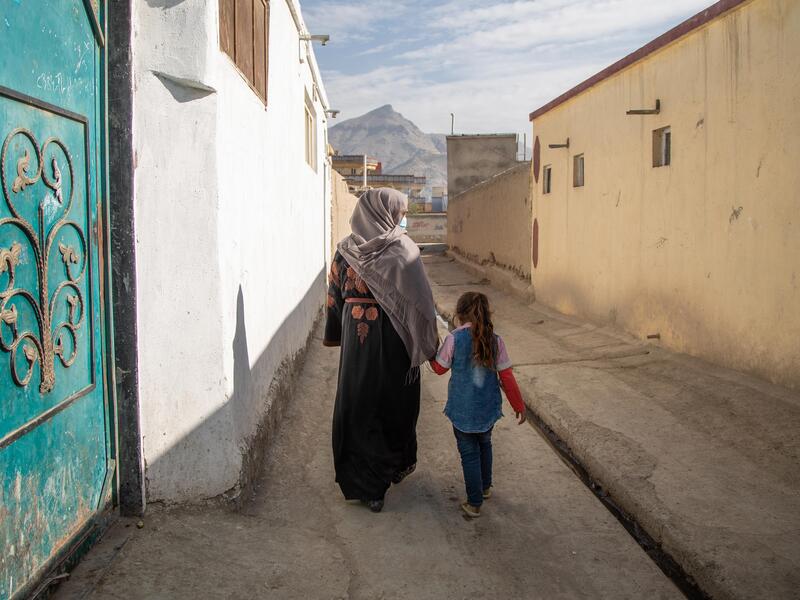 Noor and her daughter in Kabul, Afghanistan, after being forced to flee their home. With their backs to the camera, Noor and her daughter walk between two buildings holding hands. There are mountains in the distance.