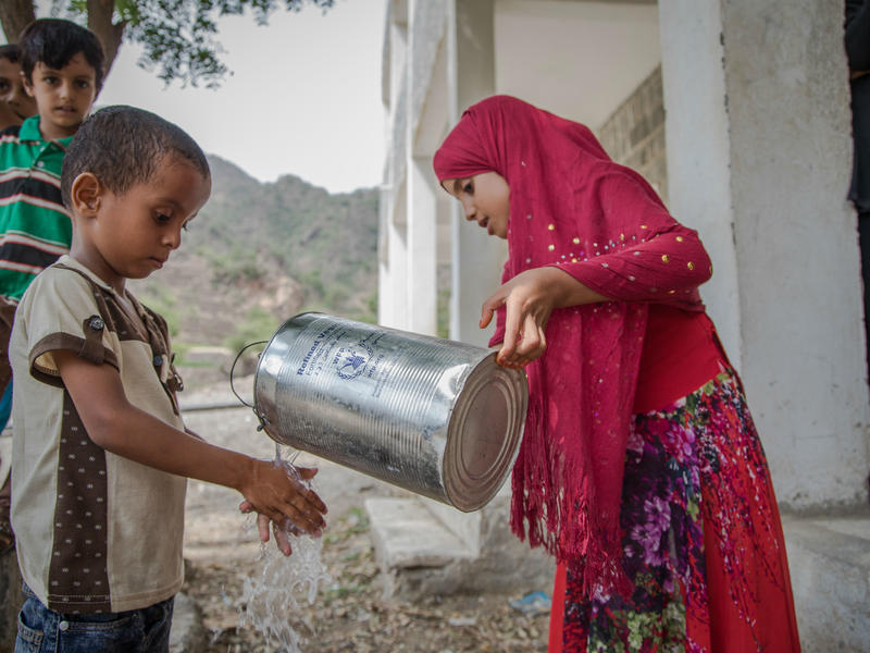 A girl  holding a jug pours water over a boy's hands so that he can wash them.