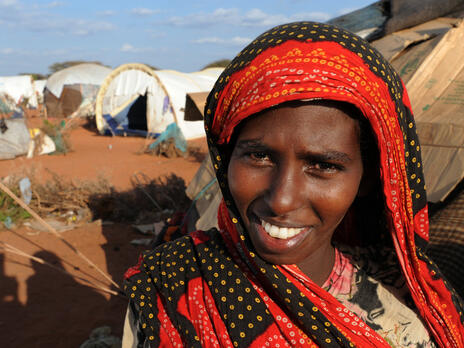 A smiling young woman stands outside a tent in Dadaab refugee camp in Kenya on a sunny day,