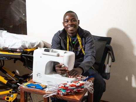 Jonathan Amissa sits at a desk with the sewing tools he uses to make masks. There is a white sewing machine and masks of all colors on the desk. He is looking at the camera and smiling. 