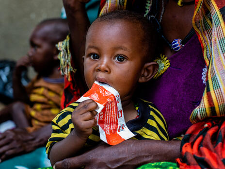 A child in Chad with acute malnutrition sits on his mother's lap at an IRC clinic, eating therapeutic peanut paste from a packet.