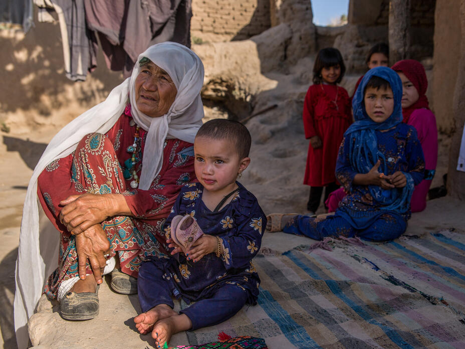In Afghanistan, a grandmother sits on the ground with a grandchild sitting next to her. There are other children sitting behind them. 
