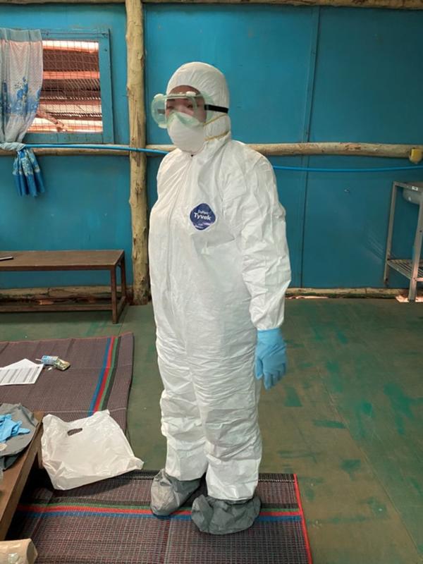 A refugee medic in Umpiem Mai camp practicing safe donning of personal protective equipment (PPE). A refugee medic stands inside in a room. They are wearing full personal protective equipment, or PPE, including a white suit, blue gloves, and goggles.