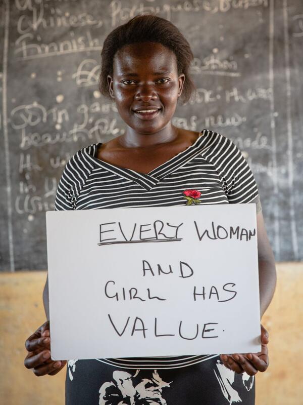 Jackie Letaru, wearing a black and white striped shirt and standing in front of a chalkboard, holds a sign that says "Every woman and girl has value." 