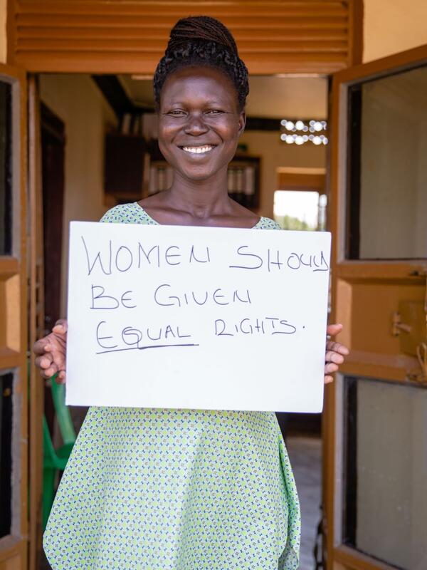 Grace Muuduru, smiling and looking at the camera, holds a sign that says "Women should be given equal rights"